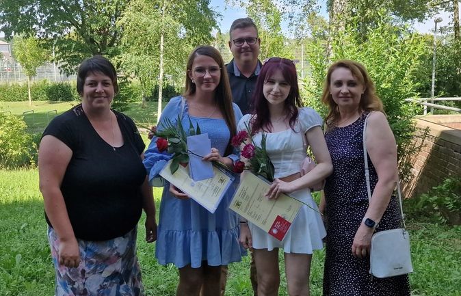 Zwei Absolventinnen mit Urkunden und Rosen zwischen Angehörigen im Freien