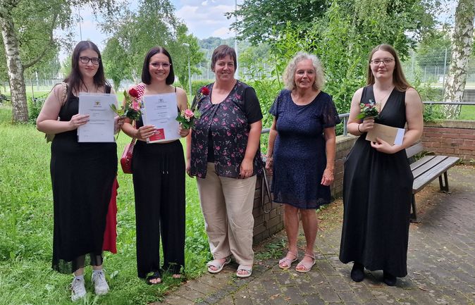 Drei Absolventinnen mit Urkunden und Blumen neben zwei Frauen
