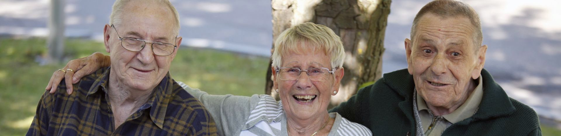 Drei ältere Personen auf Parkbank, Frau lacht, sie halten Händchen