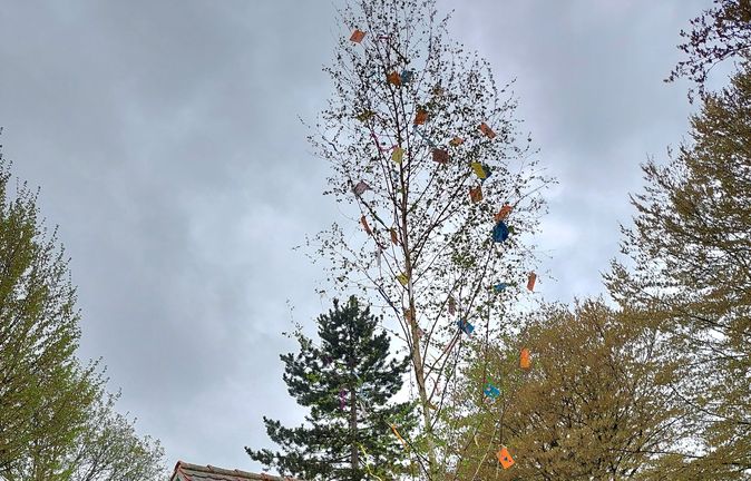 Geschmückter Maibaum mit bunten Bändern neben kleinem Häuschen